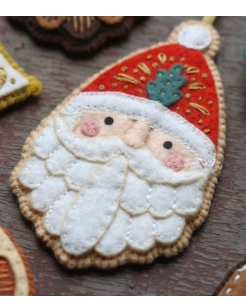 A close-up of a handmade felt Christmas ornament shaped like Santa's face, featuring a red hat with gold embroidered details, a white beard, and a small green holly accent, displayed on a dark wooden surface.