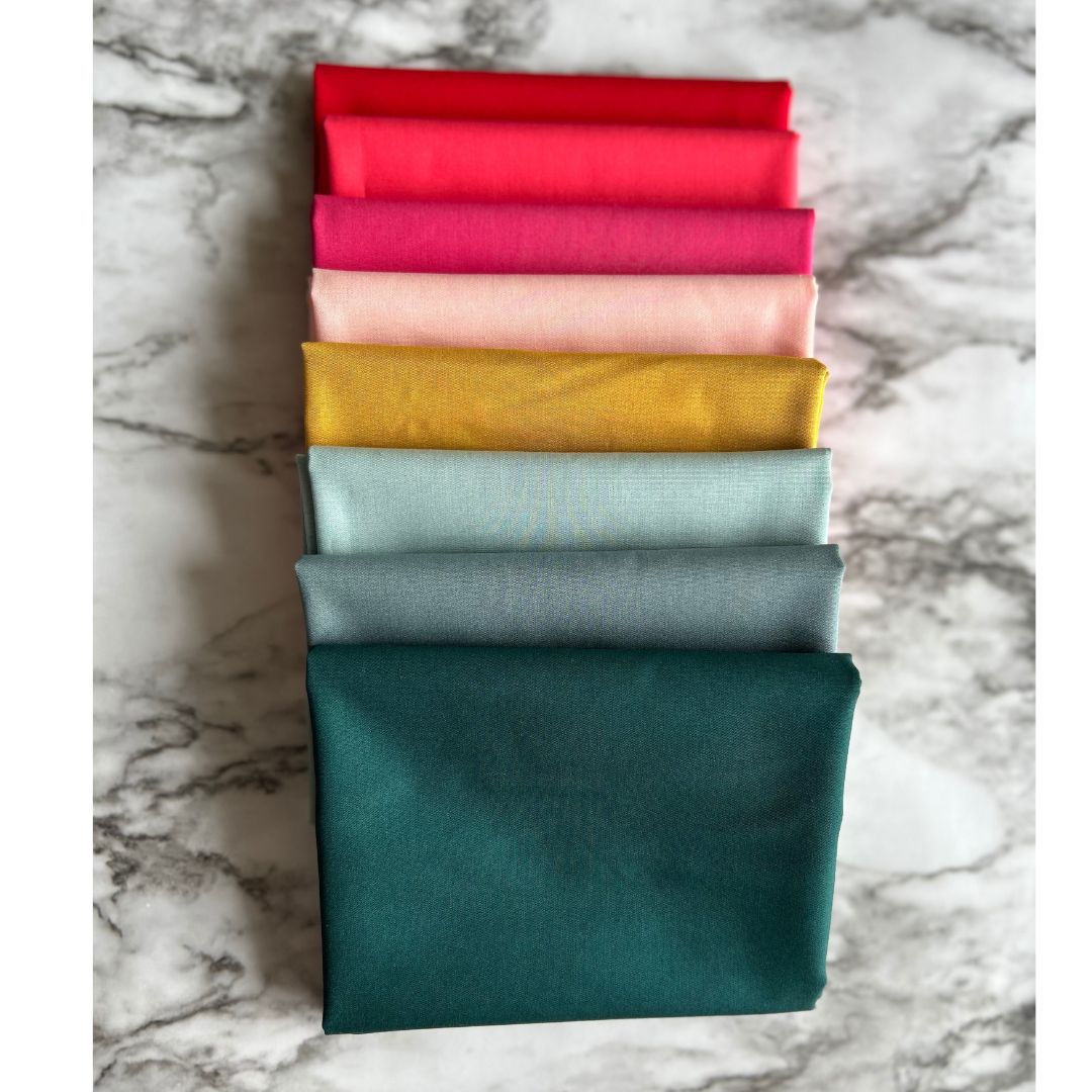 A stack of eight fat quarter pieces of fabric in various solid colors including green, blue, gray, yellow, pink, red, and orange.