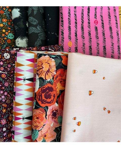 A collection of folded Halloween patterned fabrics arranged side by side, including floral prints, geometric designs, and a light pink fabric with small candy corn motifs.