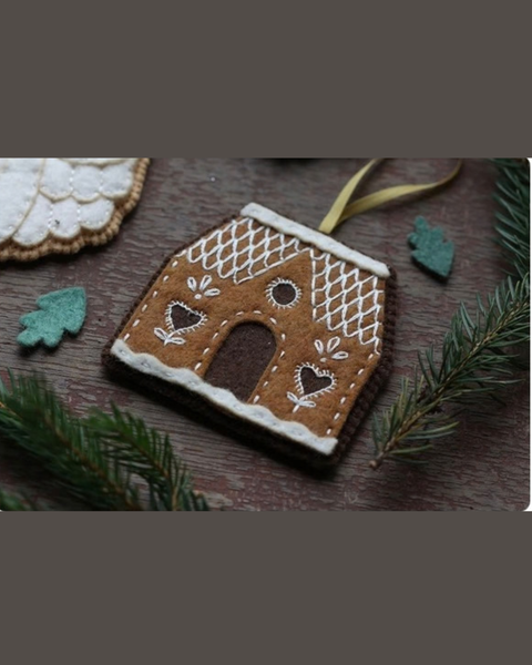A gingerbread house felt ornament with white icing-style embroidery and a gold hanging ribbon, displayed on a rustic wooden surface with evergreen branches and small green leaves.