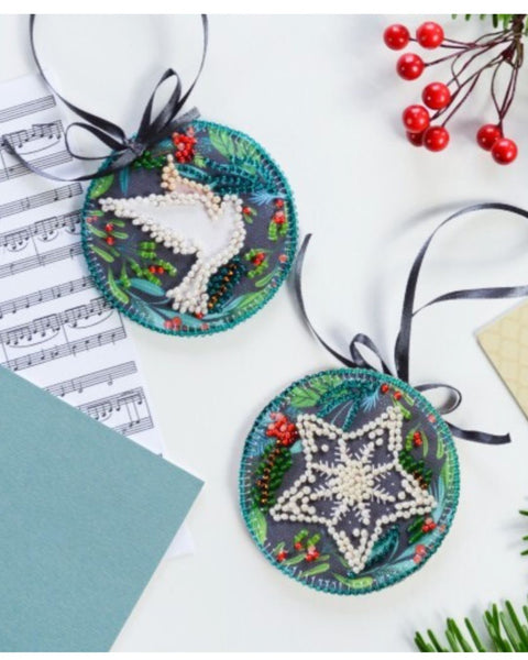 Two embroidered Christmas ornaments with a deer and star design on a white surface with music sheets and red berries.