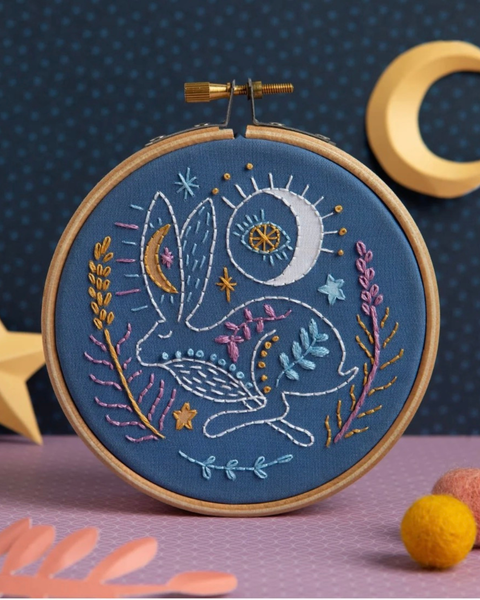 A circular embroidery hoop holding blue fabric with a whimsical stitched rabbit, moon, sun, stars, and floral motifs in white, yellow, pink, and light blue. The hoop is photographed on a pink surface against a dark blue dotted backdrop, with decorative props including a wooden crescent moon, star, and pom-poms.