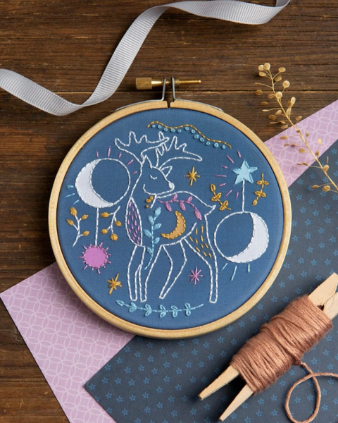 A circular embroidery hoop containing a blue fabric background with a whimsical embroidered deer, stars, moons, and floral motifs in white, yellow, pink, and teal. The hoop is styled on a wooden surface with a white ribbon, patterned paper, thread, and dried botanicals.