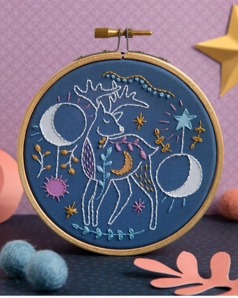 A circular embroidery hoop holding a blue fabric panel with a whimsical stitched celestial deer design in white, blue, pink, yellow, and gold tones, photographed on a styled purple background with decorative props.