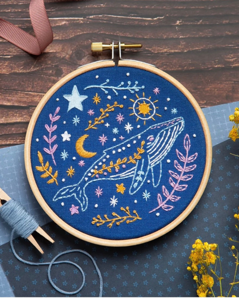 A completed circular embroidery piece in a wooden hoop, featuring a blue whale under a crescent moon and stars on a dark blue background, styled on a wooden tabletop with thread, ribbon, and small yellow flowers.