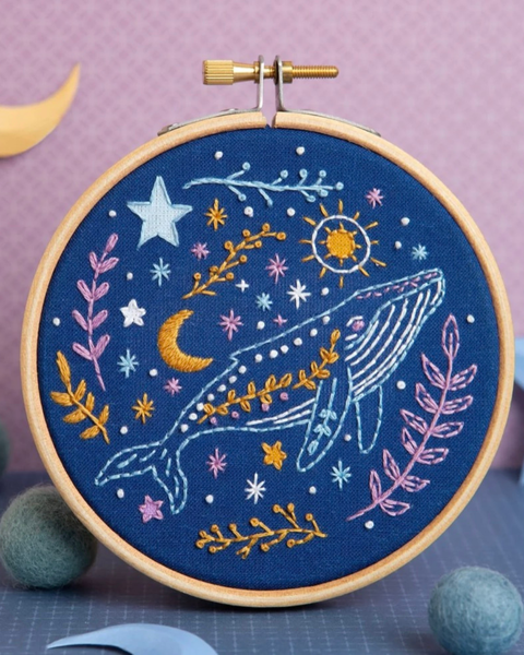 A circular embroidery hoop holding navy blue fabric with a whimsical stitched design of a whale, stars, moon, sun, and floral branches in pastel colors, photographed on a purple surface with decorative props.