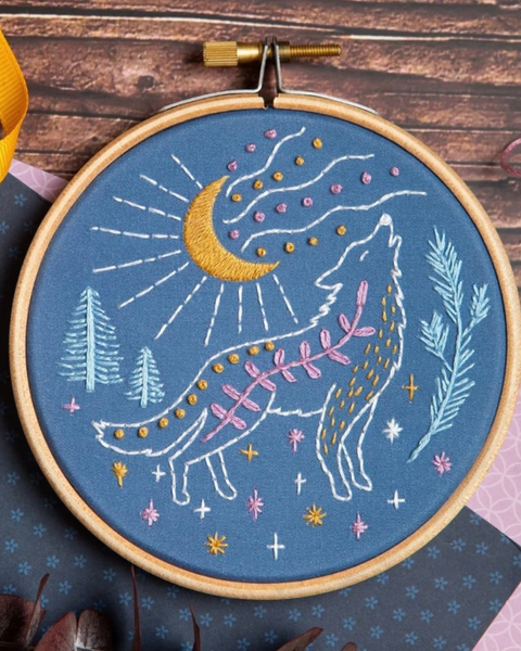 A circular embroidery hoop containing a completed embroidered design of a howling wolf under a crescent moon, with pine trees and decorative stars on a blue fabric background, photographed on a rustic wood surface.