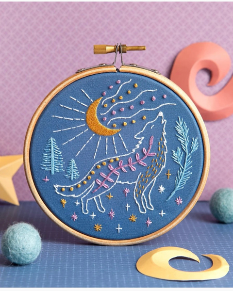 A circular embroidery hoop containing a blue fabric panel with a stitched fantasy scene of a howling white wolf under a gold crescent moon, surrounded by pine trees and stars. The hoop is displayed on a blue surface with decorative props including a gold crescent cutout, blue felt balls, and a pink decorative object against a purple patterned background.