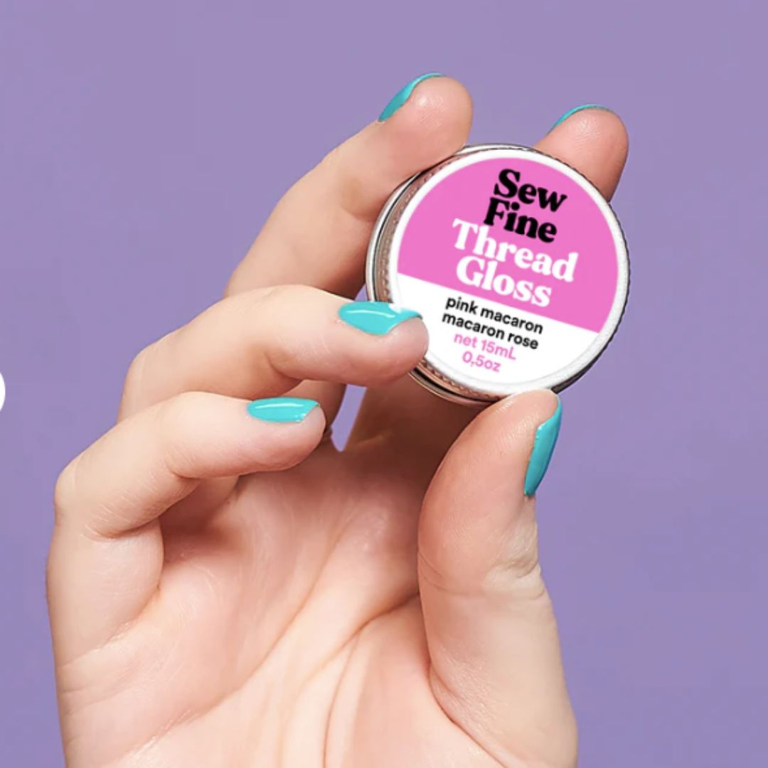 Hand holding a small container of 'Sew Fine Thread Gloss' against a purple background