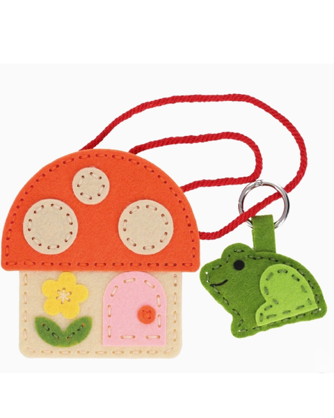 A felt mushroom-shaped keychain or bag charm with a red rope loop and a green frog-shaped ring, featuring embroidered details including a yellow flower, beige spots, and a pink door on a white background.