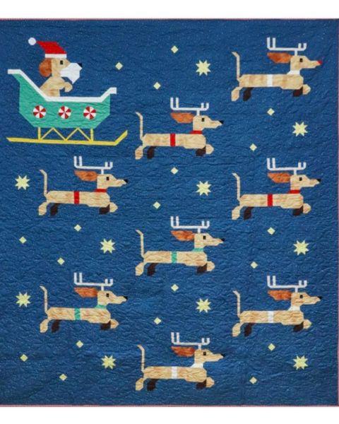 A square quilt or fabric panel featuring a blue background with a repeating pattern of tan dachshund dogs wearing red scarves and antlers, plus a Santa-clad dog in a teal sleigh and small yellow star motifs.