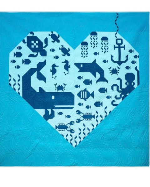A quilted textile featuring a large heart-shaped patchwork design filled with navy blue ocean-themed motifs like a whale, shark, octopus, anchor, jellyfish, and small fish on a white background, set against a bright turquoise blue quilted field.